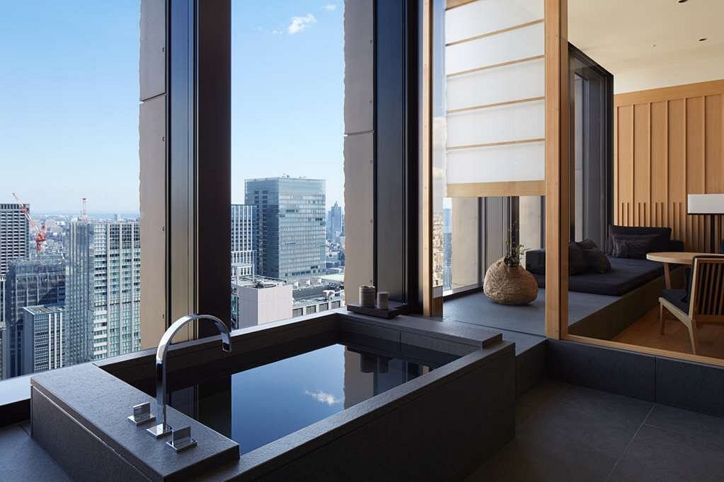 15 Unique Hotels in Tokyo: Coolest hotels 27 A modern bathroom in one of the most unique hotels in Tokyo features a deep soaking tub by large windows, revealing panoramic city skyline views; an adjacent lounge area showcases wood accents and minimalist decor.