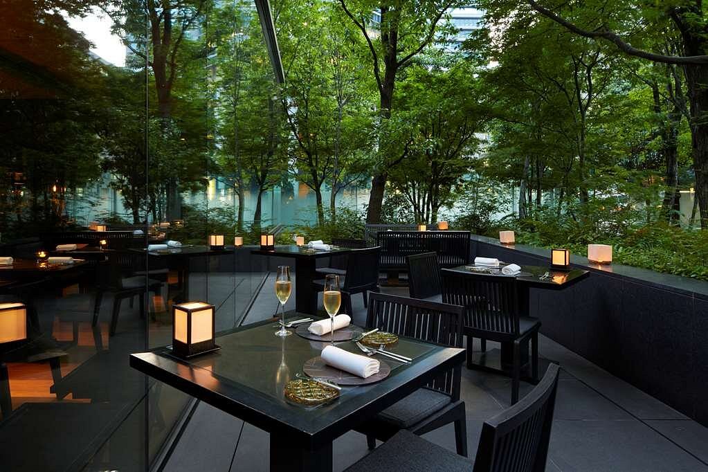 15 Unique Hotels in Tokyo: Coolest hotels 29 Outdoor dining area with black tables and chairs, set with glassware and napkins. Soft lanterns provide warm light. Surrounded by lush green trees and large windows, this space rivals those at unique hotels in Tokyo for tranquil ambiance.