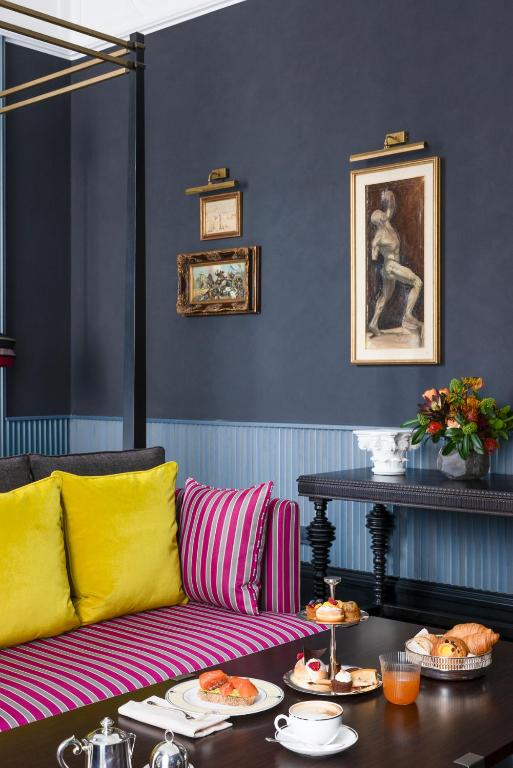 13 UNIQUE Boutique Hotels in Rome 52 A stylish living room inspired by boutique hotels in Rome, featuring a dark wall, striped pink sofa with bright yellow pillows, a black table set for tea and pastries, and artwork above a console table with fresh flowers.