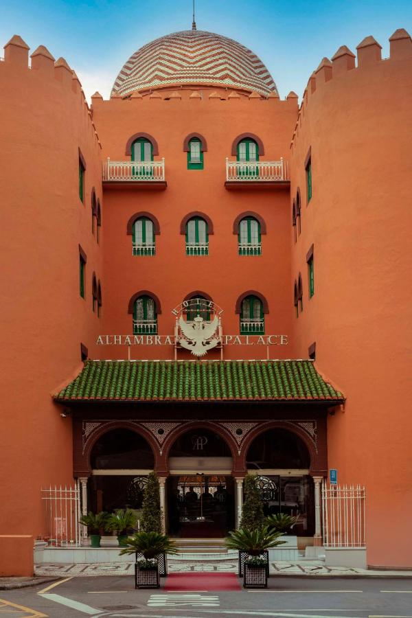 Facade of Alhambra Palace hotel, one of the renowned boutique hotels in Granada, with orange walls, green windows, arched doors, white balconies, a patterned dome, and two round towers. Plants and a canopy mark the entrance.