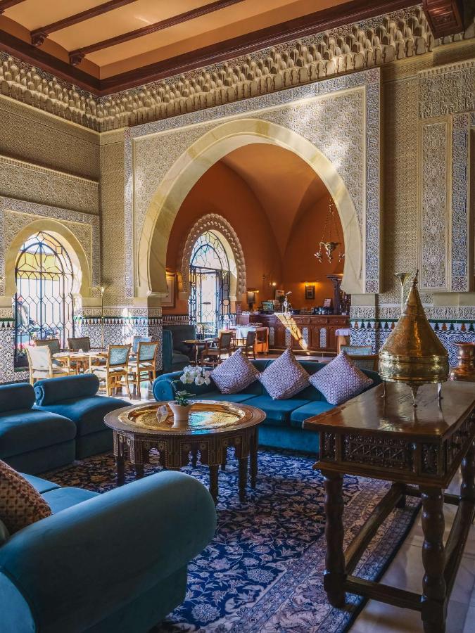 Lavish lounge with blue sofas, ornate gold tables, patterned rugs, and large arched windows. Intricate wall carvings and geometric tiles evoke the elegant, Moroccan-inspired decor found in boutique hotels in Granada.