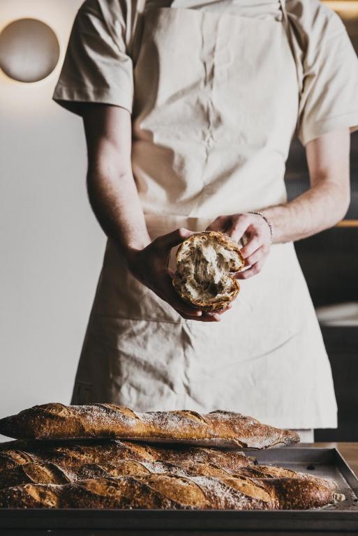 A person in a light apron breaks a loaf of rustic bread in half over a tray of baguettes, evoking the artisanal breakfasts often found at boutique hotels in Athens. The softly lit background is out of focus.