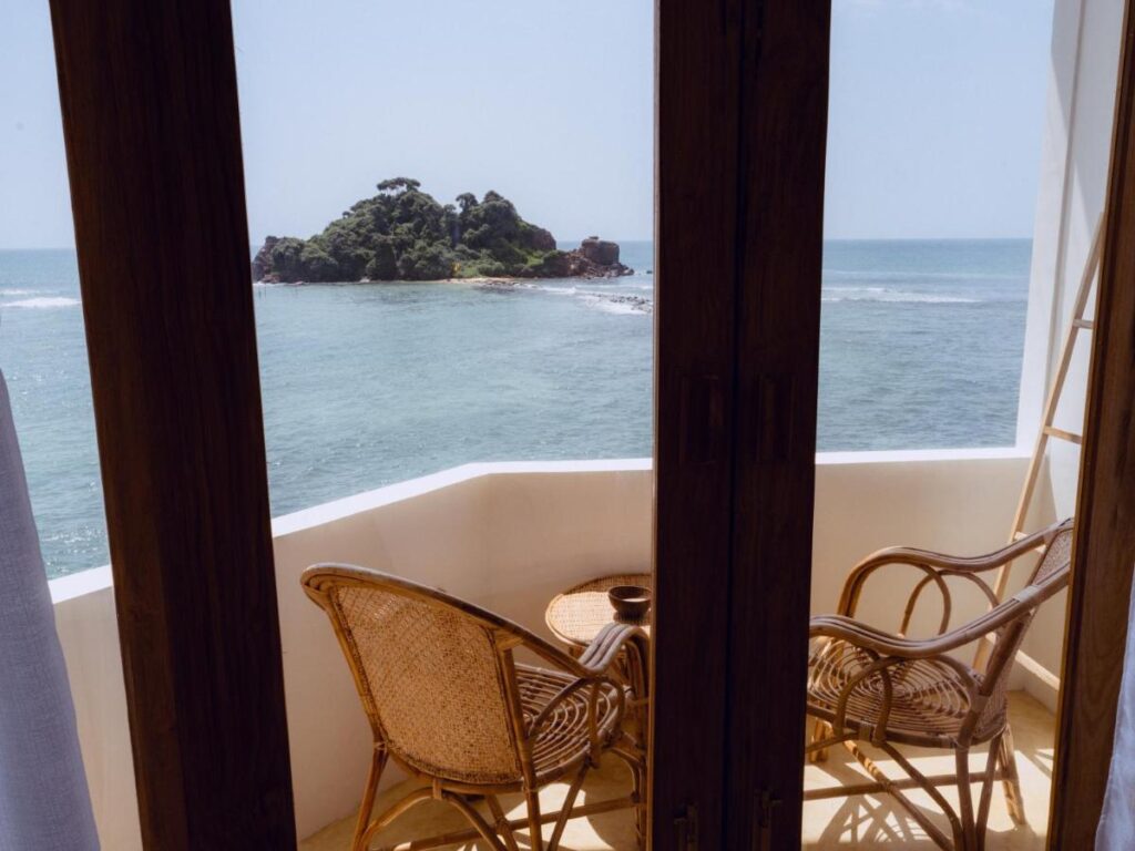 Unique Places to Stay in Sri Lanka: Boutique & Beach Hotels 3 A balcony with two wicker chairs and a small table overlooks the ocean and a tree-covered island, seen through open wooden doors—one of the unique places to stay in Sri Lanka.