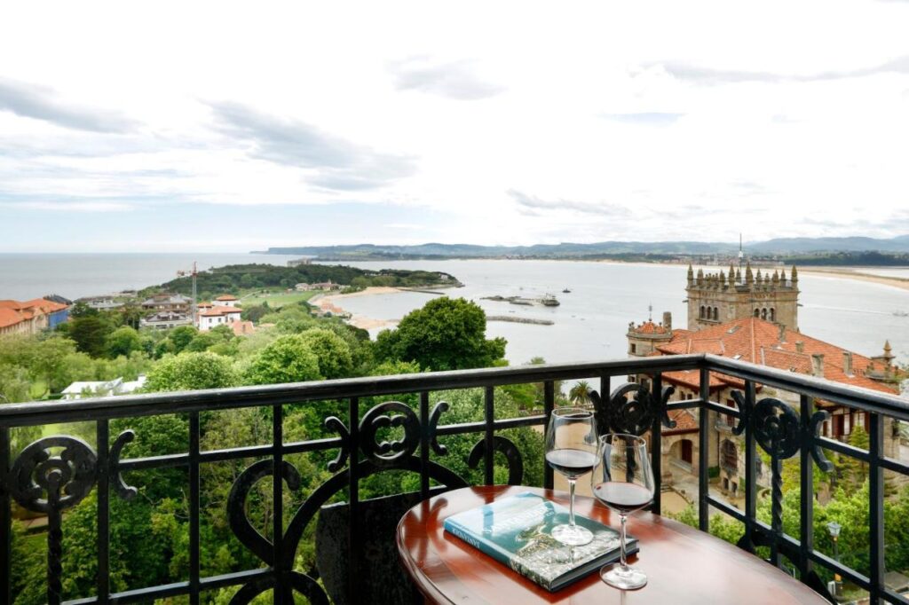 A balcony with a decorative black railing overlooks a scenic coastal town, a church, and the sea—evoking the charm of hotels in Cantabria. On the table are two glasses of red wine and an open book, with lush greenery and hills in the distance.