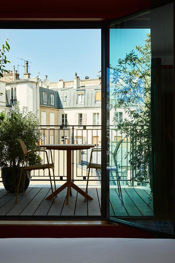 13 Unique Hotels in Paris for a Stylish Stay 18 View from inside a room at one of the unique hotels in Paris, looking out through an open glass door onto a balcony with a round table, two chairs, potted plants, and rooftops of neighboring buildings under a clear blue sky.