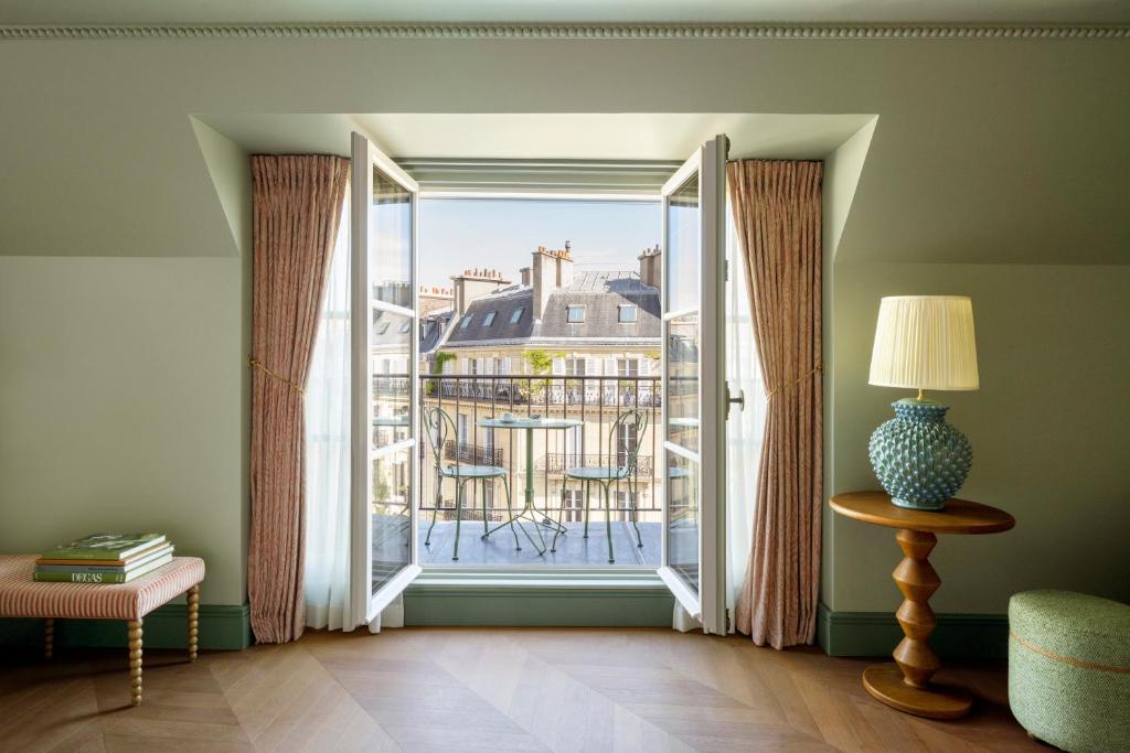13 Unique Hotels in Paris for a Stylish Stay 40 A cozy room in one of the unique hotels in Paris features pink curtains framing an open French window, revealing a small balcony with two chairs and a table. Beyond, classic Parisian rooftops beckon; a lamp sits on a round wooden table.