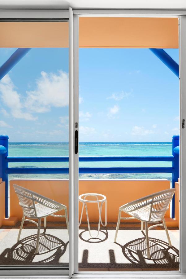 Most Unique Adults Only Hotels in Mauritius, Indian Ocean 8 Two white chairs and a small round table on a balcony of one of the finest adults only hotels in Mauritius overlook a bright blue ocean under a clear sky, seen through a glass sliding door with bold blue railings and orange walls.