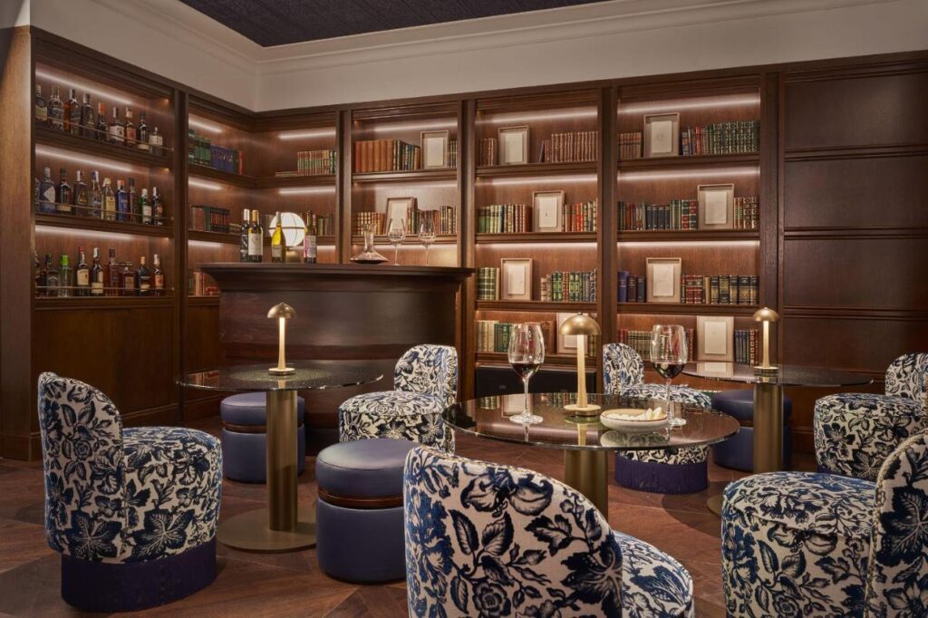 Elegant lounge with round tables, blue-patterned chairs, and a small bar—perfect for those seeking boutique hotels in Granada. Shelves lined with books and bottles adorn wood-paneled walls, while warm lighting and candles create a cozy, sophisticated atmosphere.
