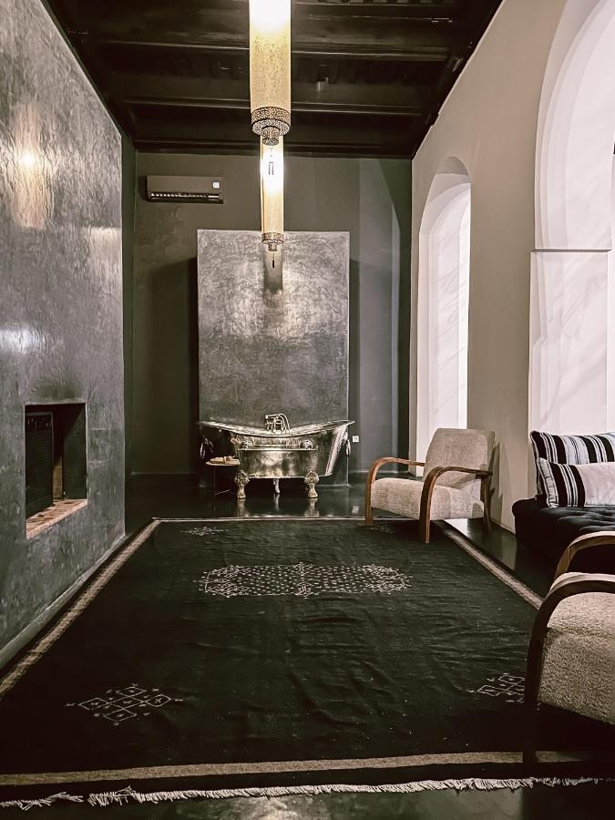 10 Unique Riads in Marrakech with Pool 30 A modern, minimalist bathroom echoes the elegance of riads in Marrakech with pool, featuring a metallic bathtub against a textured gray wall, a large black rug, two armchairs, arched windows, and a hanging pendant light.