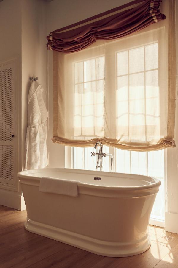 Unique Boutique Hotels in Menorca 9 A white freestanding bathtub with a towel draped over the side sits in front of a large window with sheer curtains and a brown valance, evoking the tranquil ambiance found in boutique hotels in Menorca. A white bathrobe hangs on the softly lit bathroom wall.