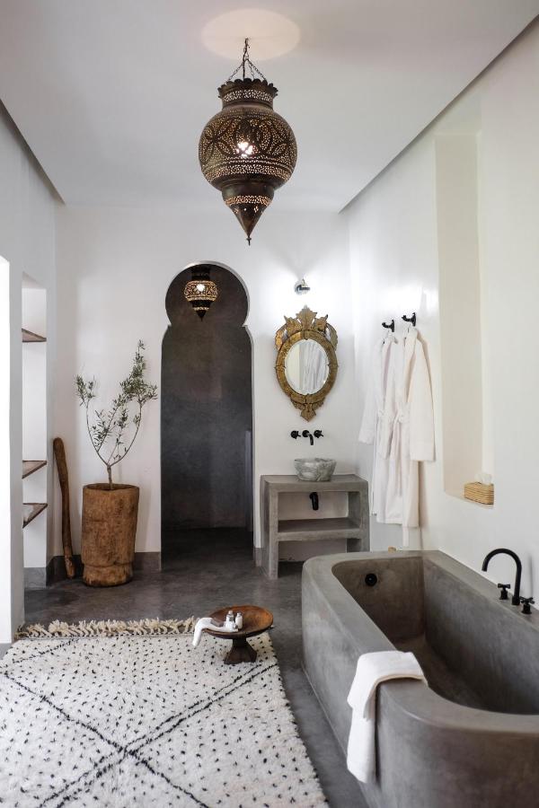 10 Unique Riads in Marrakech with Pool 33 A modern bathroom with Moroccan-inspired decor evokes the elegance of riads in Marrakech with pool, featuring a concrete bathtub, patterned rug, ornate hanging lamp, round mirror, two white robes, potted plant, rustic stool, and minimalist white walls.