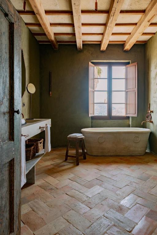 Unique Boutique Hotels in Menorca 45 Rustic bathroom reminiscent of boutique hotels in Menorca, with a herringbone tile floor, freestanding stone bathtub, wooden stool, sunlit open window, exposed ceiling beams, and a wooden vanity with baskets underneath.