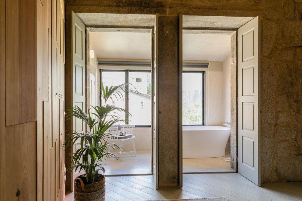 Top 12 Boutique Hotels in Porto, Portugal 21 Bright bathroom with a freestanding white tub, large windows, light walls, and a potted plant—reminiscent of the serene ambience found in boutique hotels in Porto. Natural stone doorway completes this minimalist retreat.