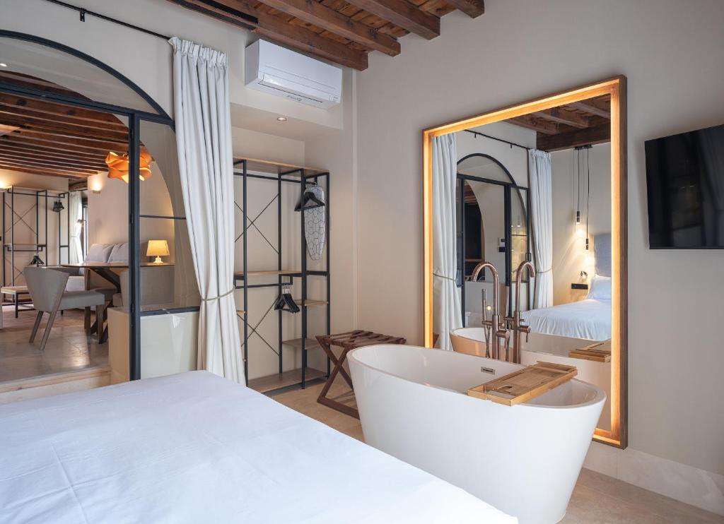 Modern hotel room in one of the finest boutique hotels in Granada, featuring a freestanding bathtub before a backlit mirror, a bed with white linens, exposed wooden beams above, and views into a cozy seating area.