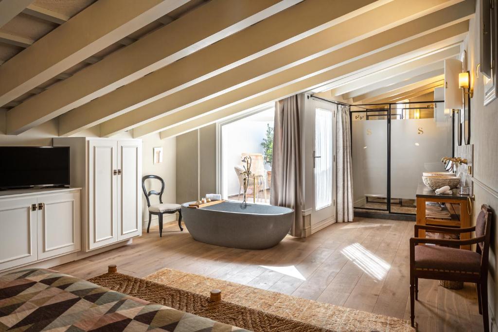 Unique Boutique Hotels in Menorca 47 A cozy attic bedroom reminiscent of boutique hotels in Menorca, with exposed beams, a freestanding bathtub, a glass-enclosed shower, a TV on a cabinet, and natural light streaming through large curtained windows onto wooden floors.