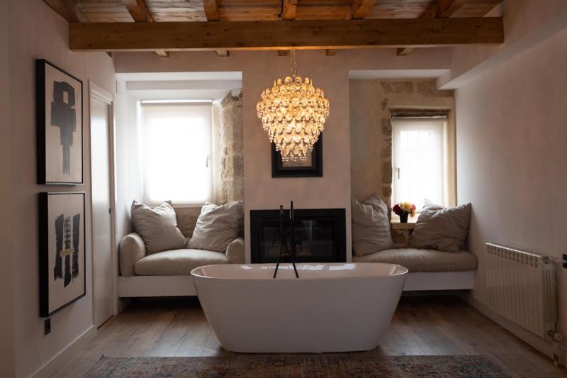 A cozy bathroom reminiscent of boutique hotels in Cantabria, with a freestanding white tub, crystal chandelier, window seats with cushions, a fireplace, wood-beamed ceiling, contemporary art on the wall, and sunlight filtering through the windows.