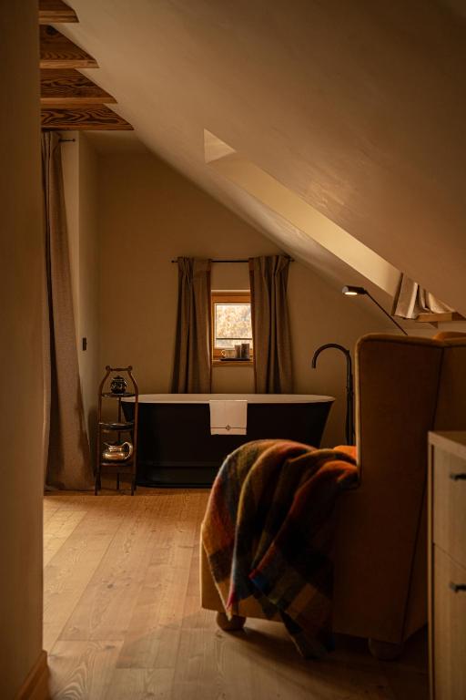 A cozy attic bathroom reminiscent of the best hotels in the Dolomites, featuring a freestanding black bathtub under a small window, beige curtains, wooden floors, and a chair draped with a colorful plaid blanket in warm lighting.