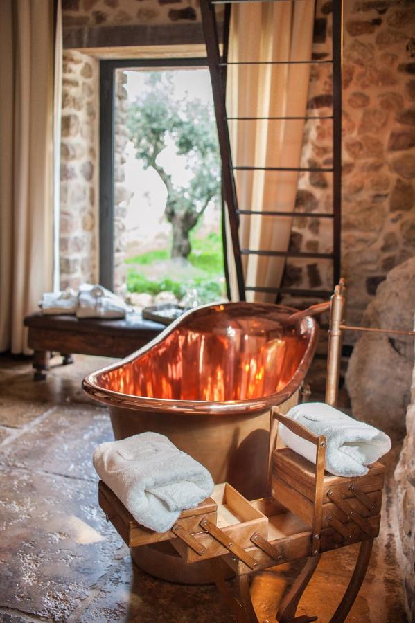 TOP 10 Luxury Hotels in Andalucía 5 A rustic bathroom, inspired by luxury hotels in Andalucia, features a shiny copper bathtub, a wooden towel rack with neatly folded towels, stone walls, a large window framing a tree outside, and soft natural light filling the space.