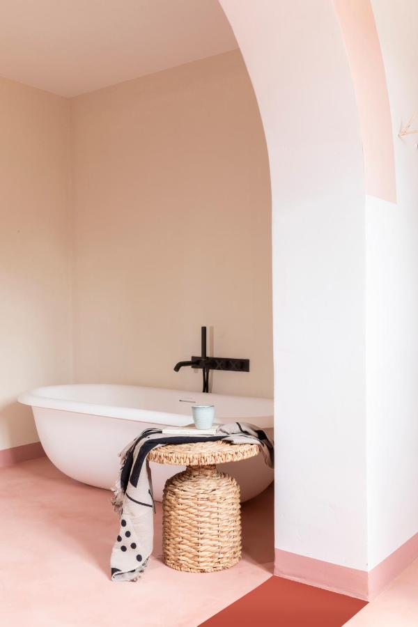 Unique Boutique Hotels in Menorca 23 Minimalist bathroom with a white freestanding tub, black faucet, and a woven stool holding a cup and towel. Soft pink floors evoke the calm, modern vibe found in boutique hotels in Menorca, complemented by an elegant arched entryway.