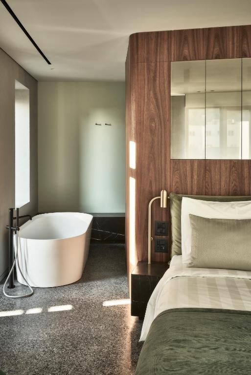 Modern bedroom with green and white bedding, a wooden accent wall, and a gold lamp on the nightstand. An open bathroom area features a white freestanding bathtub. The space is filled with natural light, echoing the elegant style of boutique hotels in Athens.