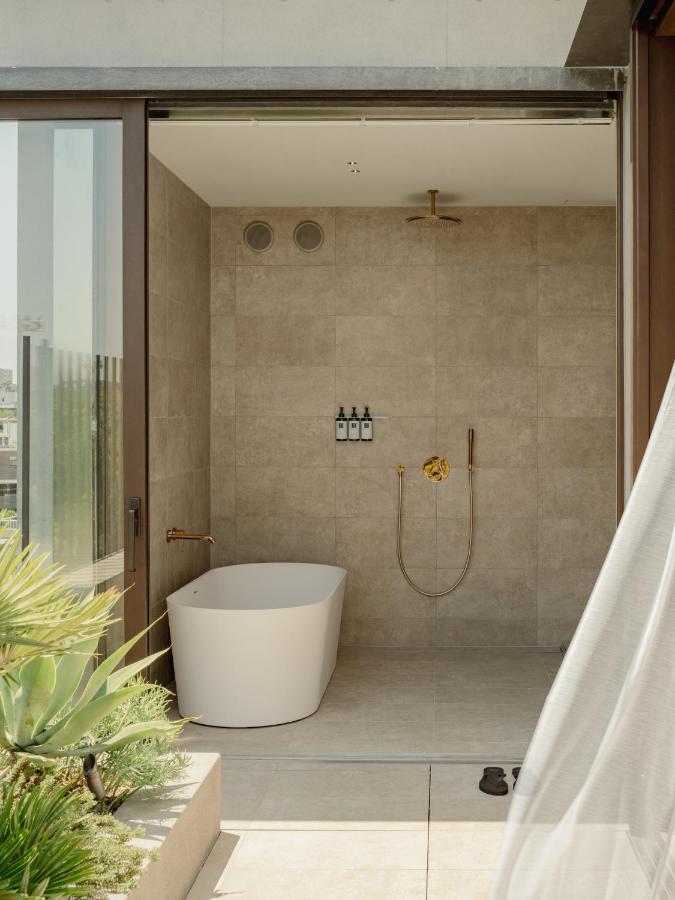 15 Unique Hotels in Tokyo: Coolest hotels 5 A modern outdoor bathroom at one of the unique hotels in Tokyo, featuring beige tile walls, a freestanding white bathtub, gold fixtures, and three bottles on a shelf. Sunlight streams through the open glass door beside lush green plants.
