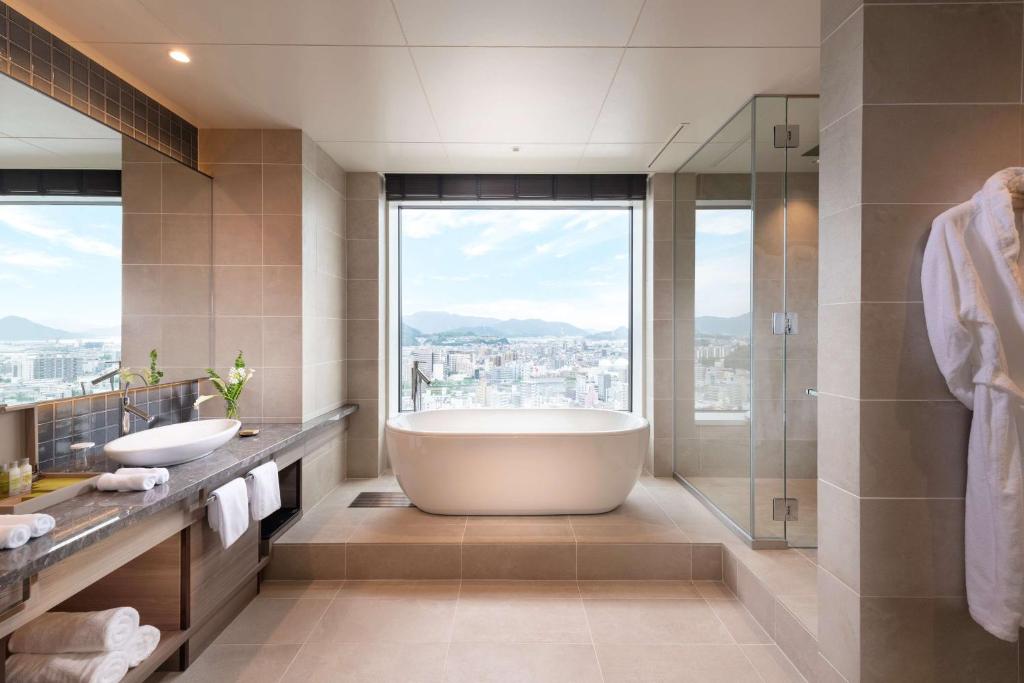 7 Best Hotels in Hiroshima, Japan 21 Modern bathroom at one of the best hotels in Hiroshima, featuring a freestanding bathtub by a large window with cityscape and mountain views, double sinks on the left, and a glass shower with a white bathrobe on the right.