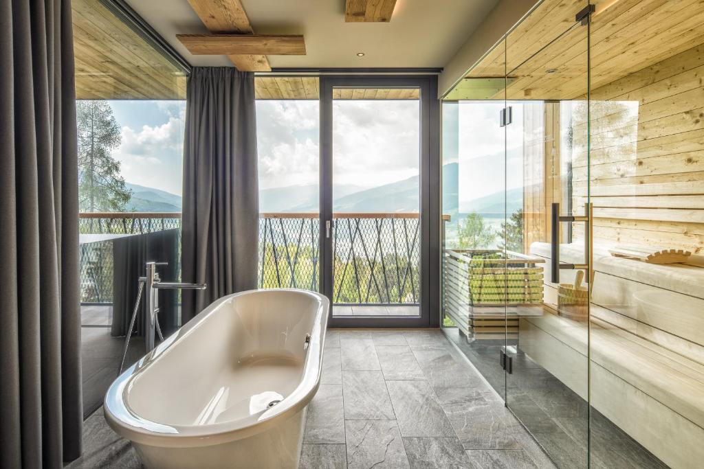 Modern bathroom with a freestanding white bathtub, large windows framing mountain and lake views, dark curtains, and a glass-enclosed wooden sauna—reminiscent of wellness spaces in the best hotels in the Dolomites.