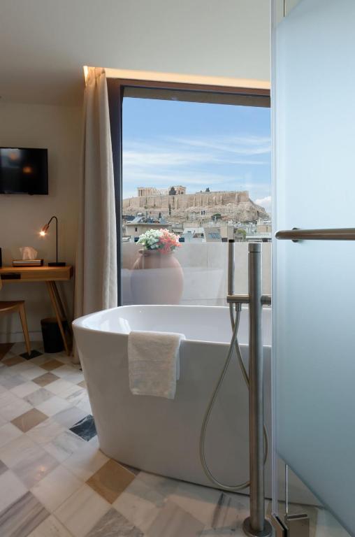 Modern bathroom with a white freestanding bathtub, towel hanging on the side, and large window offering a clear view of the Acropolis—an elegant touch often found in boutique hotels in Athens. Desk, lamp, and TV are visible in the background.