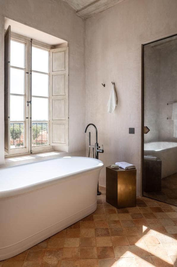 Unique Boutique Hotels in Menorca 4 A minimalist bathroom inspired by boutique hotels in Menorca features a white freestanding bathtub, large window with wooden shutters, beige textured walls, a gold side table, and warm terracotta tile flooring bathed in natural light.