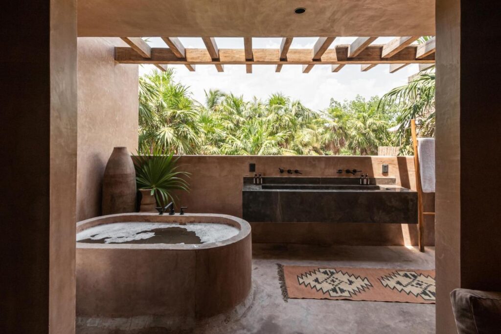 Top Hotels in Tulum, Mexico: Luxury and Beachfront Stays 6 A rustic outdoor bathroom inspired by the top hotels in Tulum, with a soaking tub, double sink, large vases, and potted plants. Wooden beams line the ceiling; lush palm trees sway beyond the open wall and a patterned rug adorns the floor.