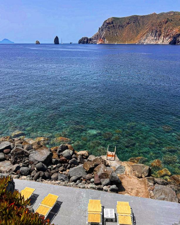 7 Best Hotels in the Aeolian Islands 20 Clear blue sea with a rocky shore, yellow lounge chairs on a patio at one of the charming hotels in the Aeolian Islands, and a ladder leading into the water. Rocky cliffs and small islands appear in the distance under a bright blue sky.