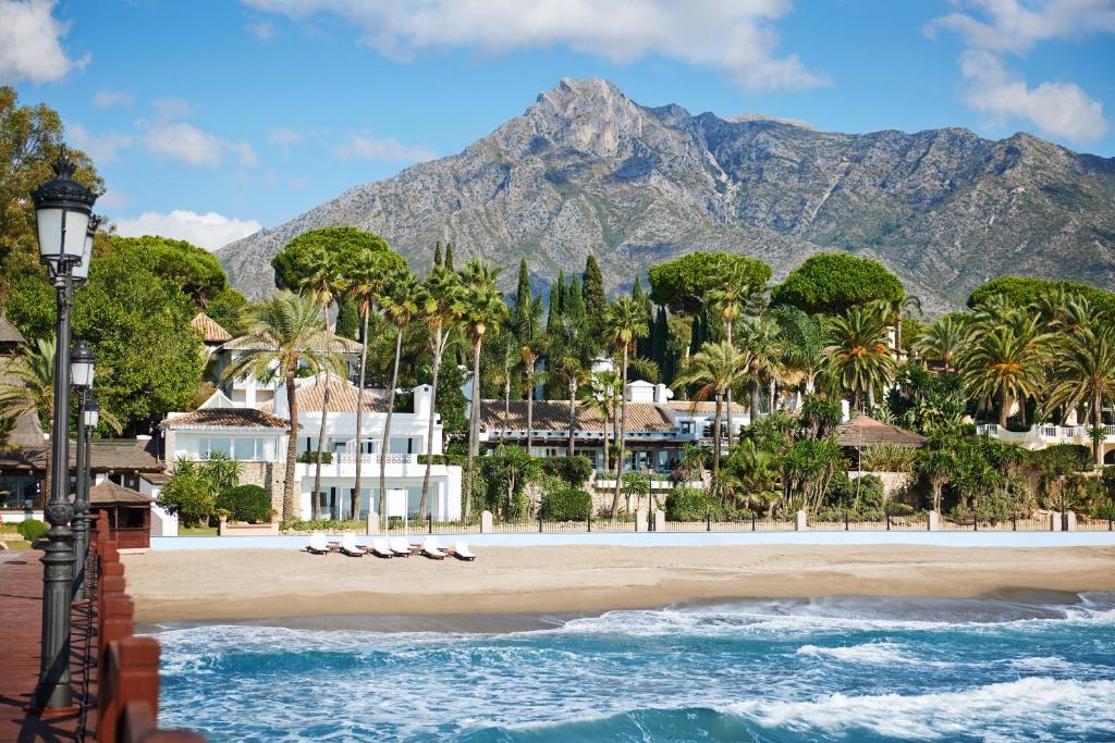 TOP 10 Luxury Hotels in Andalucía 19 A sandy beach with gentle waves, lined with lounge chairs and palm trees, sits in front of luxury hotels in Andalucía. In the background, a mountain range rises under a blue sky with scattered clouds.