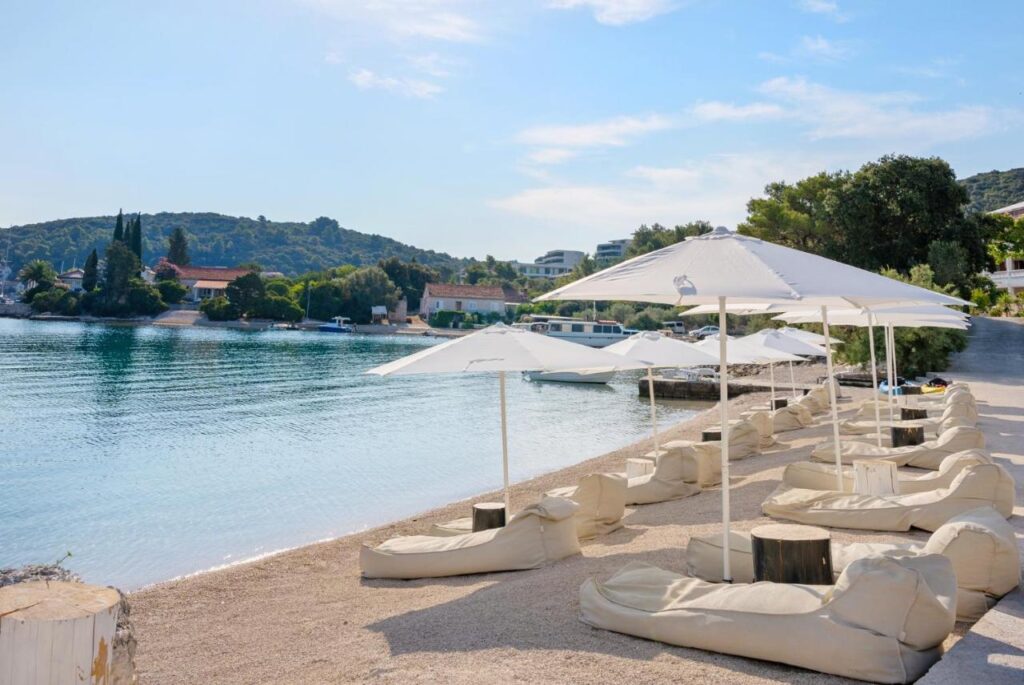 Boutique Hotels in Korčula Island 4 A calm beach with white umbrellas and cushioned lounge chairs on sandy shore, overlooking clear blue water and green hills—near boutique hotels in Korcula—with houses and trees in the background under a partly cloudy sky.