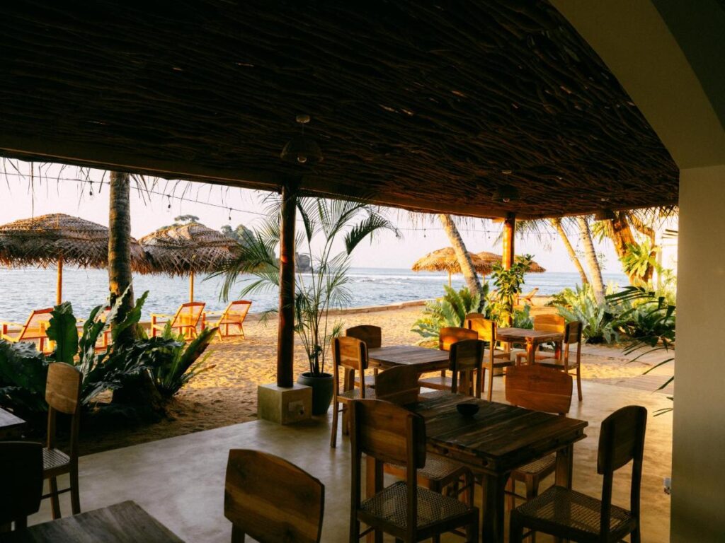 Unique Places to Stay in Sri Lanka: Boutique & Beach Hotels 4 Open-air beachside restaurant with wooden tables and chairs, palm trees, and thatched umbrellas facing the ocean. This relaxing tropical scene is perfect for travelers seeking unique places to stay in Sri Lanka right by the water.