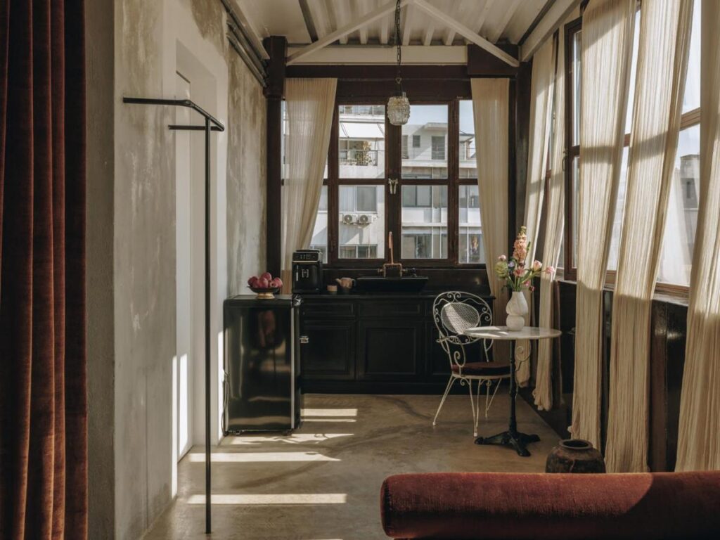 Sunlit room with large windows, sheer curtains, a small black kitchen area, black mini fridge, iron chair at a round table with flowers, and minimalist clothes rack against concrete walls—like the style found in the best boutique hotels in Athens.
