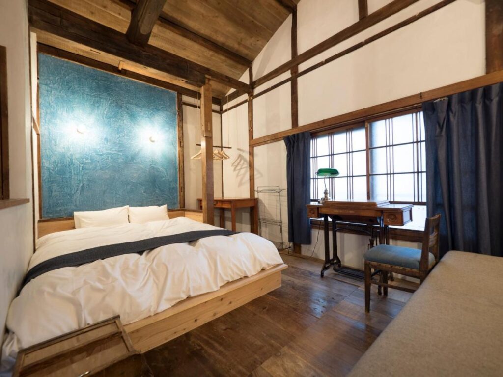 15 Unique Hotels in Tokyo: Coolest hotels 50 A cozy bedroom with a wooden floor and ceiling, a double bed with white bedding, blue accent wall, and dark blue curtains—perfect for those seeking the charm of unique hotels in Tokyo. A desk with a green lamp sits invitingly by the window.