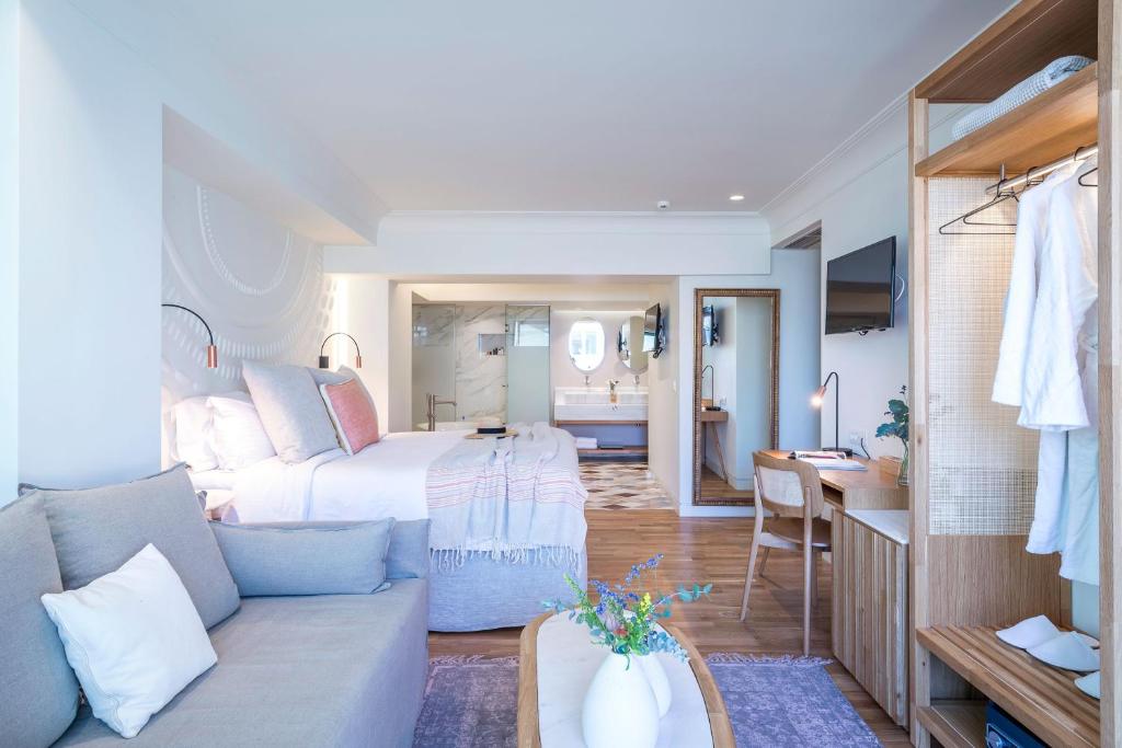 Modern hotel room with a large bed, sofa, desk, open wardrobe, and bathroom with glass shower. Bright and decorated in neutral tones, it features wooden furnishings—capturing the stylish comfort of boutique hotels in Athens.