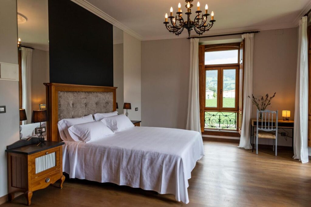 Bright bedroom with wooden floors, a large bed with white bedding, two nightstands with lamps, a chandelier, and a window overlooking greenery—perfect for those seeking hotels in Cantabria with serene views. A desk and chair sit by the window with a small plant.