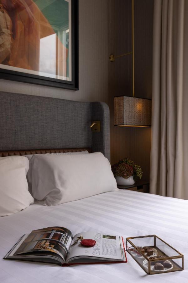 13 UNIQUE Boutique Hotels in Rome 4 A neatly made bed with white linens, often seen in boutique hotels in Rome, Italy, sits beside a nightstand holding a small bouquet and a lamp. An open magazine and a tray with jewelry rest on the bed, while framed artwork hangs above the headboard.