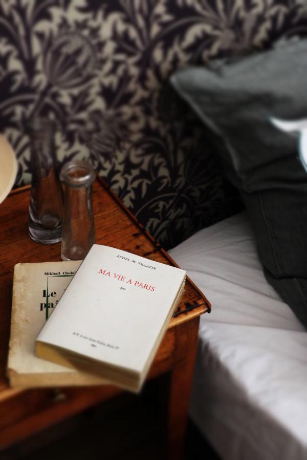 13 Unique Hotels in Paris for a Stylish Stay 15 A wooden bedside table in one of the unique hotels in Paris holds two books, including Ma Vie à Paris, beside an empty glass bottle, drinking glass, and a bed with white sheets and a gray pillow. Patterned wallpaper decorates the background.
