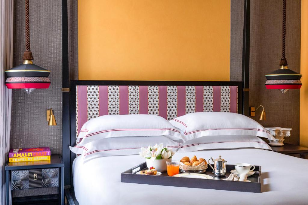 13 UNIQUE Boutique Hotels in Rome 55 A neatly made bed with white linens and a patterned headboard sits between two stylish pendant lights, evoking the charm of boutique hotels in Rome. A breakfast tray with coffee, juice, and pastries rests on the bed beside books and decor items.