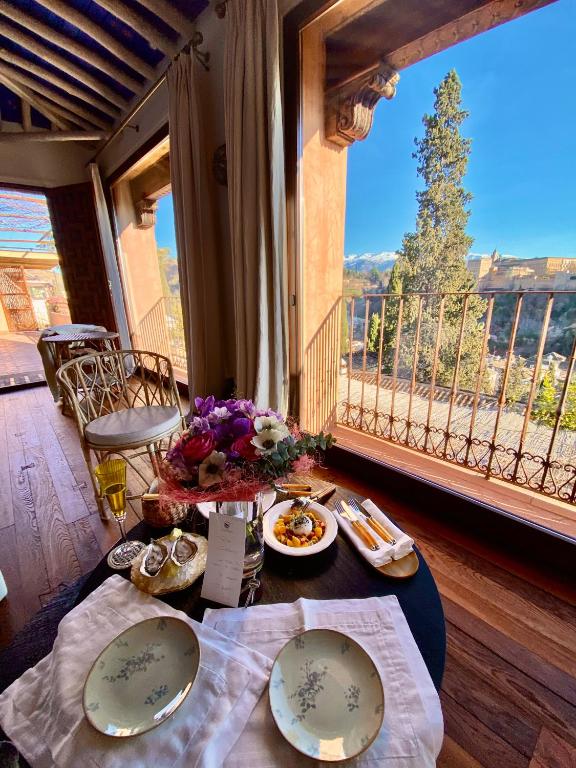 A cozy table set for two with floral plates, a bouquet, and food stands by large windows overlooking trees and distant hills recalls the inviting charm of boutique hotels in Granada, with ornate chairs and wooden floors completing the scene.