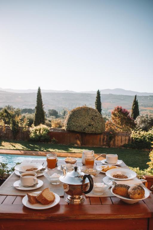 TOP 10 Luxury Hotels in Andalucía 39 A table set for breakfast outdoors at one of the luxury hotels in Andalucía features tea, juice, bread, pastries, and cereal bowls, overlooking green trees, bushes, and distant mountains beneath a clear sky.