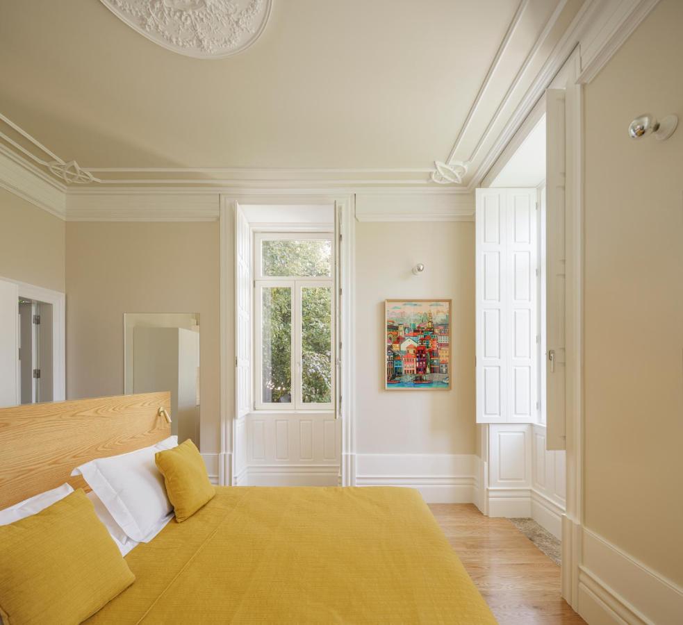 Top 12 Boutique Hotels in Porto, Portugal 12 A bright bedroom reminiscent of boutique hotels in Porto, with beige walls, a wooden bed topped with yellow bedding, white shuttered windows letting in natural light, a colorful painting on the wall, and intricate ceiling moldings.