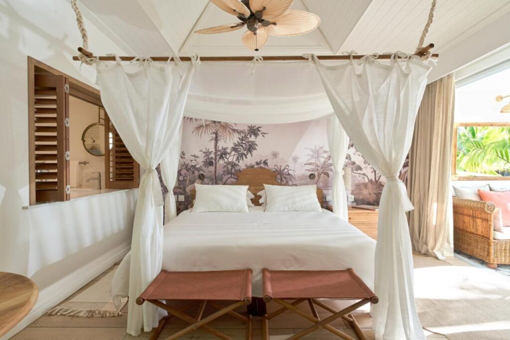 Most Unique Adults Only Hotels in Mauritius, Indian Ocean 1 A bright, airy bedroom in one of the top adults only hotels in Mauritius, with a canopy bed draped in white curtains, tropical wall art, wooden furniture, beige accents, and large windows that flood the room with natural light.