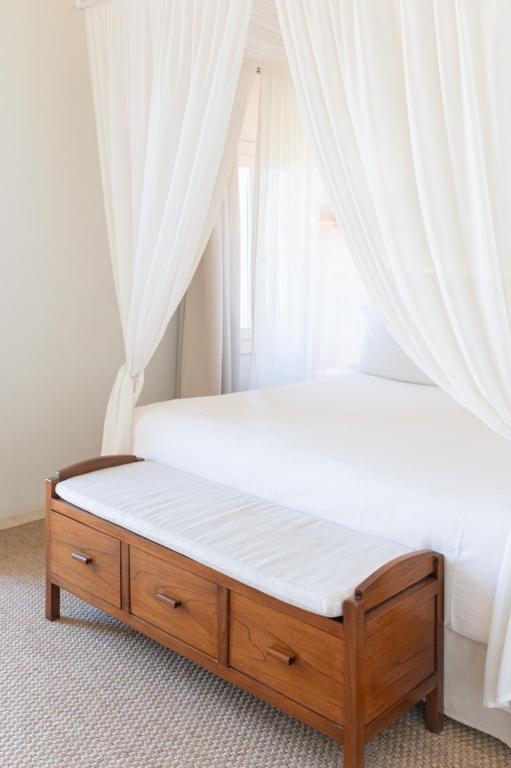 Unique Boutique Hotels in Menorca 39 A bright bedroom reminiscent of boutique hotels in Menorca, featuring a white canopy bed, sheer white curtains, and a wooden bench with drawers at the foot of the bed, all atop a light woven rug.
