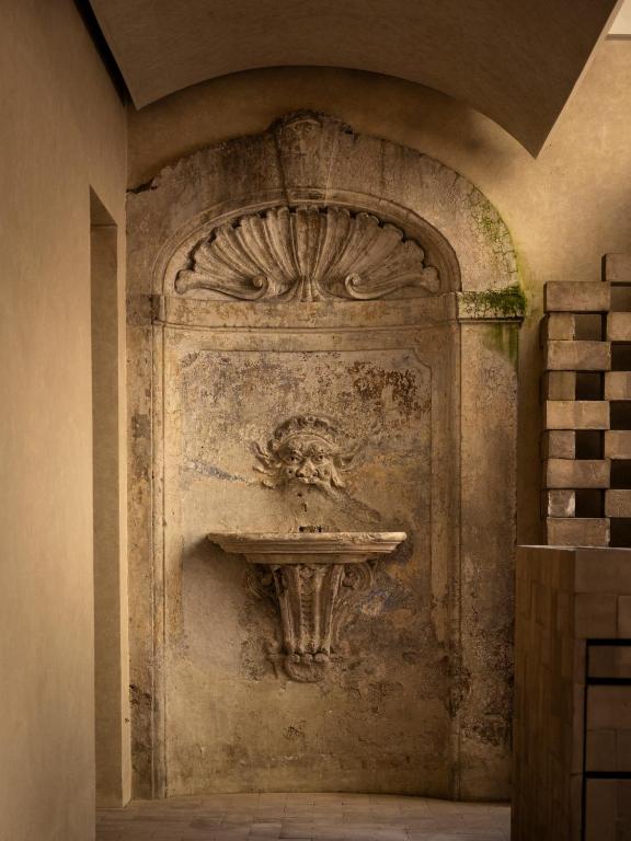 13 UNIQUE Boutique Hotels in Rome 30 A weathered stone fountain with a sculpted face and a shell motif is set into an old, textured wall—evoking the timeless elegance often found in boutique hotels in Rome. Moss and stains suggest age. Stacked stone blocks sit nearby in the softly lit corridor.
