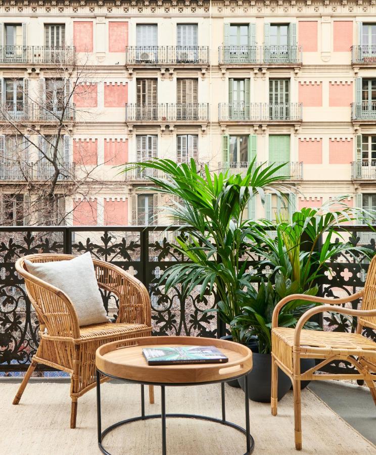 13 Unique Hotels in Barcelona, Spain 7 A cozy balcony in one of the unique hotels in Barcelona features wicker chairs, a round wooden table, potted green plants, and an ornate railing overlooking a decorative building with many windows and balconies.