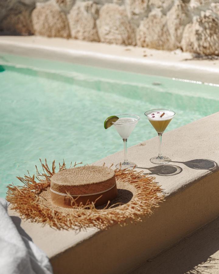 10 Unique Riads in Marrakech with Pool 18 A straw hat and two cocktails in martini glasses sit on the edge of a sunlit swimming pool, reminiscent of the tranquil vibes found at riads in Marrakech with pool, with turquoise water and a textured stone wall in the background.