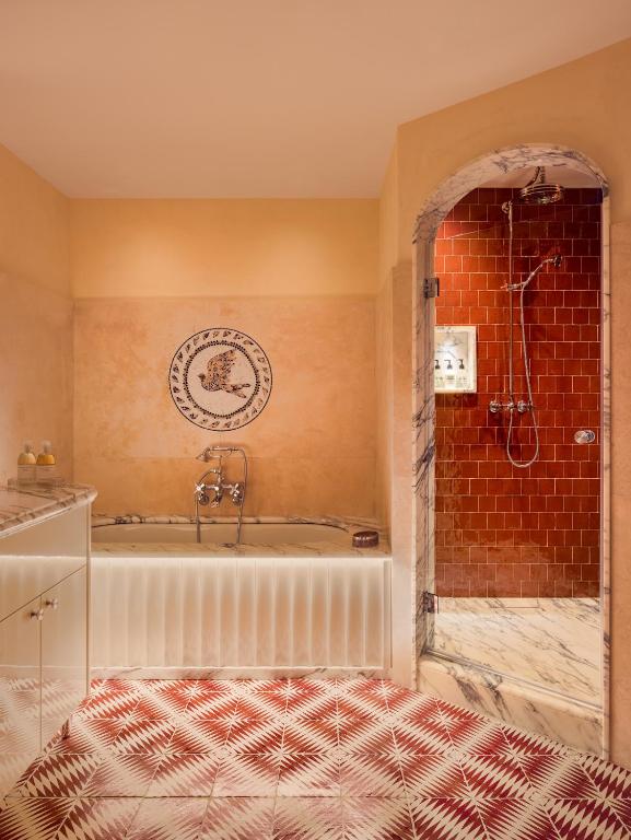 13 UNIQUE Boutique Hotels in Rome 42 Elegant bathroom reminiscent of boutique hotels in Rome, featuring a marble bathtub, mosaic bird artwork, red and white patterned floor tiles, and an arched entrance to a shower with red tiles and marble trim.